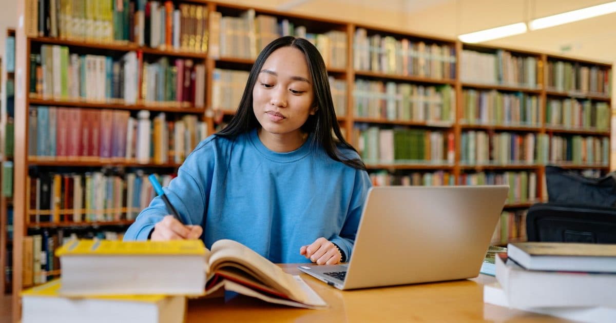 Estudiante universitaria repasando apuntes con libros y laptop en una biblioteca