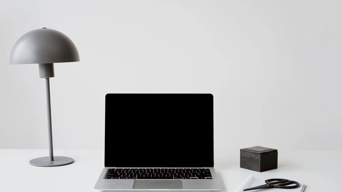 A minimalist office setup featuring a closed laptop, modern lamp, and a notepad with scissors on a white background.