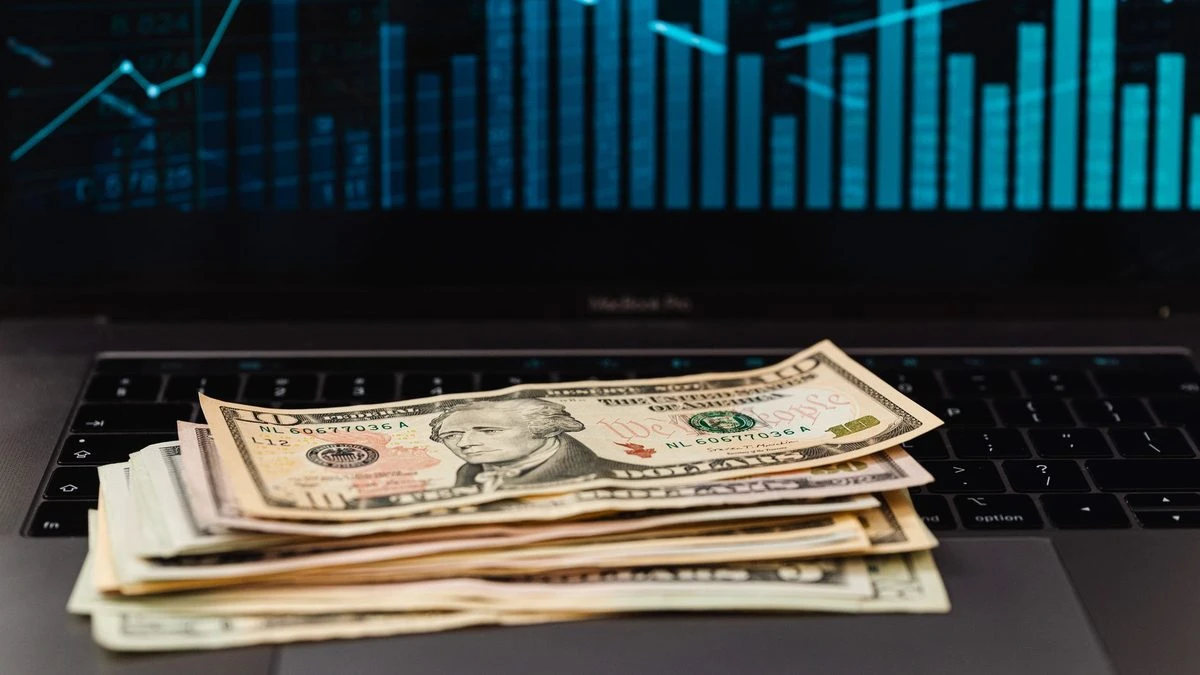 US dollar bills on laptop with financial graph in background, symbolizing wealth and technology.