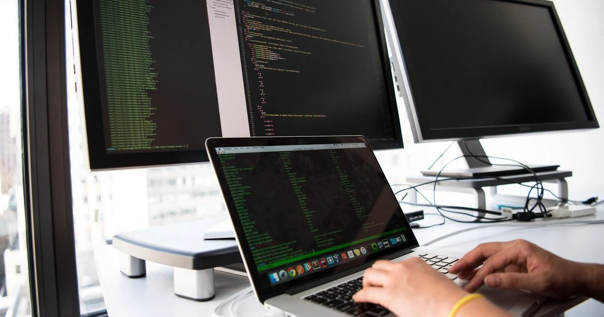 Developer typing on a laptop displaying code, with multiple monitors showing code in a bright office.