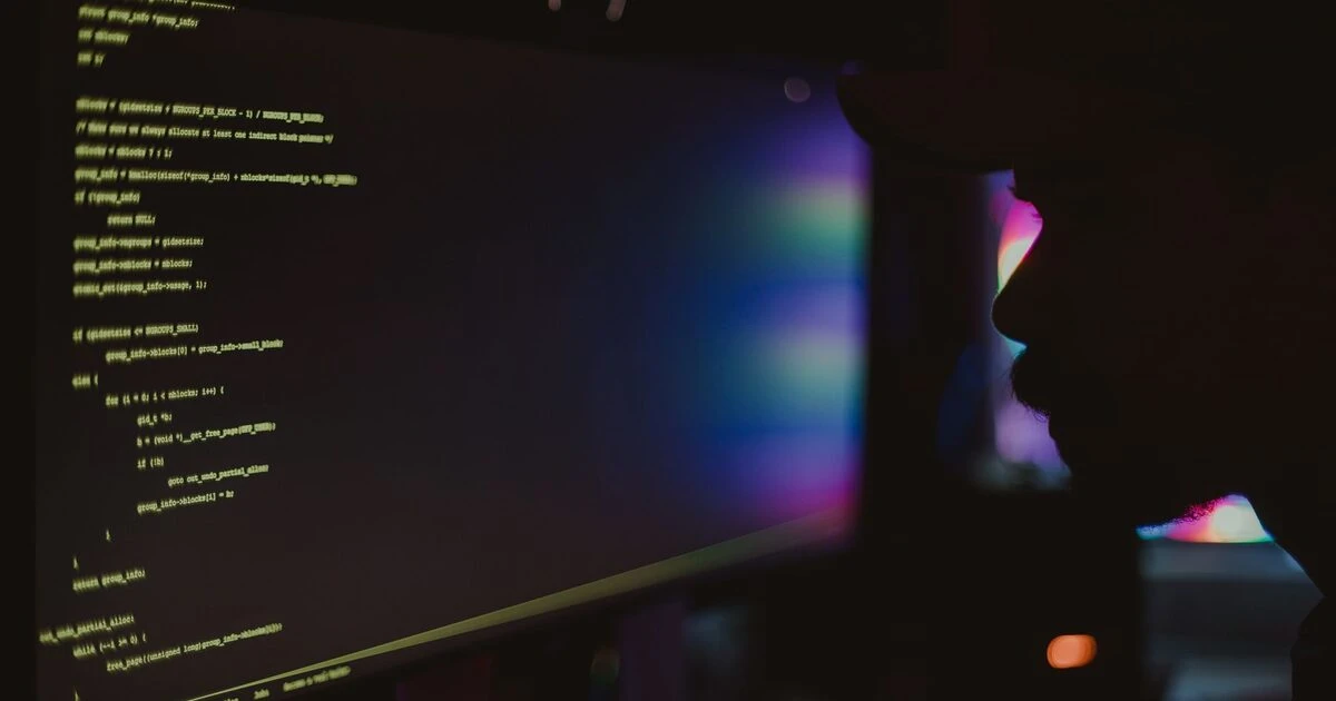 Developer reviewing code on a monitor in a dark room, silhouetted beside glowing terminal-style text.