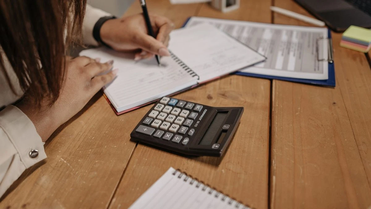 Woman writing notes with a calculator on a wooden desk, focusing on work and accounting.