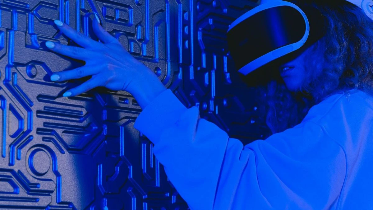 A woman wearing a VR headset touches a textured wall in a blue-lit futuristic environment.
