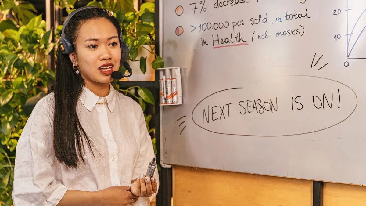 Woman presenting business statistics on whiteboard using headset, engaging audience.