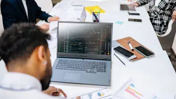 Team collaborating in an office setting, discussing financial graphs on a laptop screen.