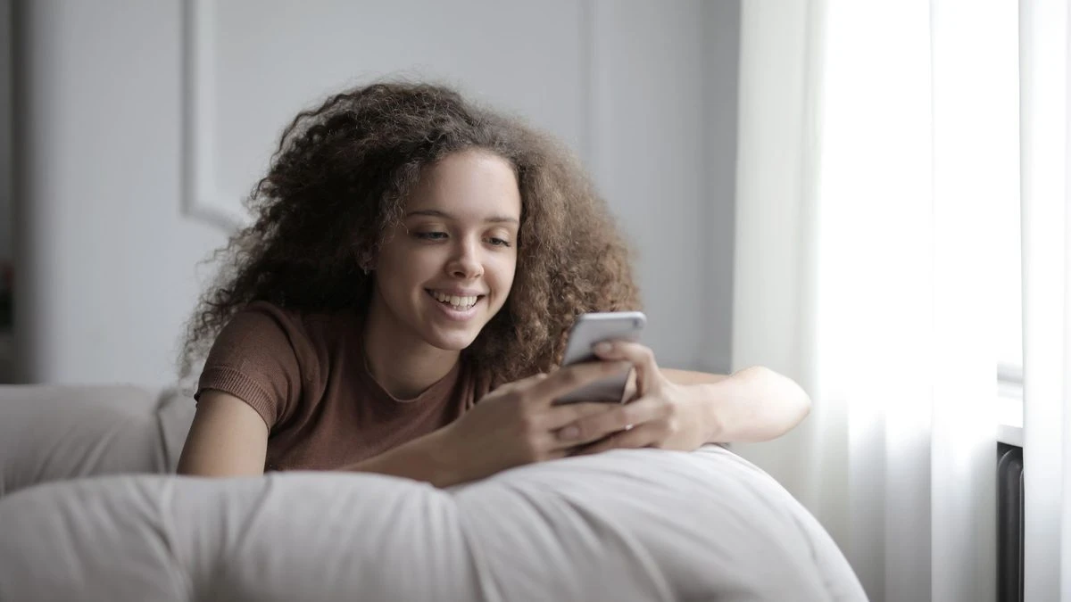 Curly teenage girl in casual clothes smiling while browsing mobile phone resting on cozy sofa in living room at home