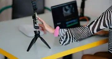 Creator setting up a smartphone on a tripod beside a laptop for producing video content.
