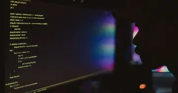 Developer reviewing code on a monitor in a dark room, silhouetted beside glowing terminal-style text.