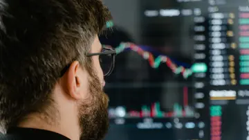 A bearded man with glasses examines trading charts on a computer screen, indicating financial analysis.