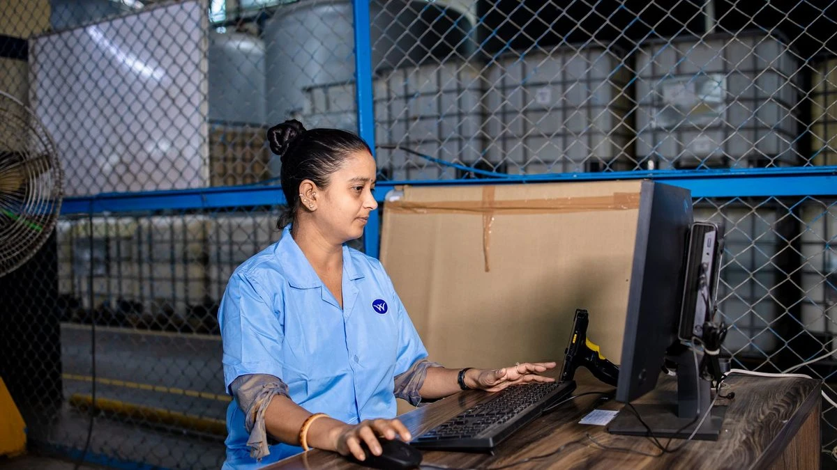 A woman in blue uniform operates a computer inside a factory warehouse, focusing on her task.